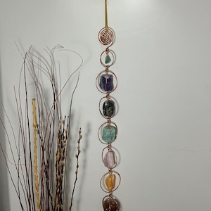 May include: A vertical wall hanging featuring a copper wire spiral at the top, followed by a series of circular copper wire frames. Each frame contains a raw, multi-colored gemstone, including green, purple, blue, light green, pink, yellow, and brown stones.