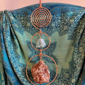 May include: A hanging ornament featuring a copper wire spiral at the top, followed by two copper wire circles. The upper circle holds a small, pale green translucent crystal, and the lower circle contains a large, textured, reddish-brown crystal.
