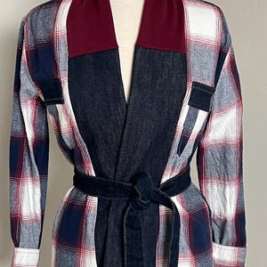 May include: A long-sleeve jacket featuring a mix of dark blue denim and a red, white, and blue plaid flannel pattern. The garment includes a solid maroon shawl collar, a matching denim tie belt at the waist, and two small denim chest pockets.