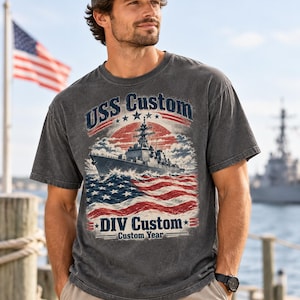 May include: A charcoal grey short-sleeve t-shirt featuring a graphic of a naval warship sailing over an American flag. The text reads USS Custom, DIV Custom, and Custom Year. The shirt is paired with a grey baseball cap with an American flag patch.