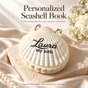 Può includere: Un libro a forma di conchiglia personalizzato con una copertina in conchiglia bianca naturale sfumata di rosa sulla parte superiore. Sulla conchiglia è stampato il testo nero 'Laura' e 'My 30th'. Due anelli in metallo argentato fissano le pagine.