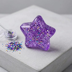 Könnte beinhalten: Ein sternförmiges Objekt aus durchscheinendem violettem Harz, gefüllt mit buntem Glitzer. Daneben befindet sich ein kleiner transparenter Behälter mit Deckel und ein Haufen loser, bunter, kreisförmiger Glitzerpartikel.