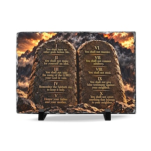 May include: A rectangular slate plaque featuring the Ten Commandments in gold text. The design shows two stone tablets set against a dramatic mountain landscape at sunset. The text lists commandments I through X clearly on the textured stone surface.
