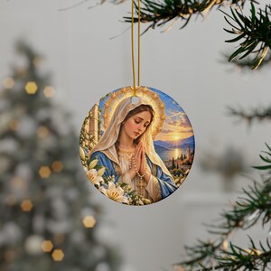 May include: A round ceramic ornament featuring a detailed illustration of a woman with a head covering, wearing blue and white robes, holding a rosary with hands clasped in prayer. A golden halo surrounds her head, with white lilies and a sunset landscape.