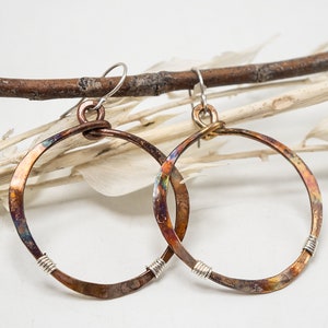 May include: A pair of handcrafted hoop earrings with a rustic, textured appearance. The earrings feature a copper-colored metal with iridescent hues of blue, purple, and gold. The bottom of each hoop is wrapped with silver wire, and they hang from silver hooks.