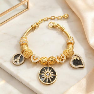 May include: A gold-tone charm bracelet featuring a snake chain with various beads and pendants. Charms include a black circular Eiffel Tower disc, a black circular starburst disc, two heart-shaped charms, and several gold filigree beads with crystals.