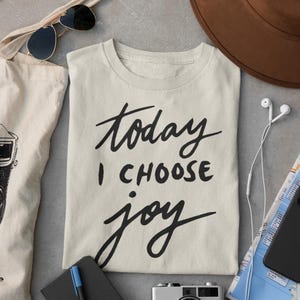 Puede incluir: Una camiseta de manga corta de color crema con el texto impreso en negro "today I CHOOSE joy" en una mezcla de letra cursiva y de imprenta. La camiseta está rodeada por un sombrero marrón, gafas de sol, una bolsa, un cuaderno, un bolígrafo, una cámara y auriculares.