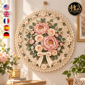 Puede incluir: Un tapiz circular de ganchillo que presenta un ramo central de flores rosas y color crema con hojas verdes. La pieza está enmarcada por un borde decorativo de color crema con pequeños motivos florales y un lazo en la parte inferior.