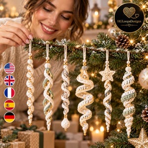 May include: Six macrame Christmas ornaments hanging from a pine branch. The ornaments feature twisted cord designs in cream, sage green, and grey, accented with gold beads and white pearls. One ornament is shaped like a star.