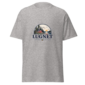 Könnte beinhalten: Ein graumeliertes T-Shirt mit einem kreisförmigen Aufdruck, der eine rote Hütte an einem See mit Tannen und einem Sonnenuntergang zeigt. Der Text lautet LUGNET SCANDINAVIAN WAY OF LIVING mit einer kleinen schwedischen Flagge darunter.