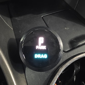 May include: A circular black automotive gear selector dial featuring a digital display. The screen shows the letter P with the word PARK in white text above the word DRAG in bright teal text, set against a dark background within the vehicle console.