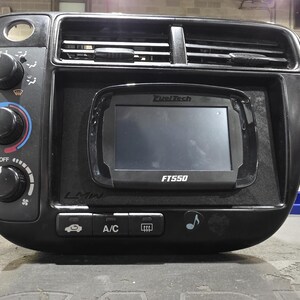Puede incluir: Un panel de consola central de tablero automotriz negro que presenta una pantalla digital FuelTech FT550, rejillas de ventilación integradas, tres perillas de control circulares y una fila de botones con símbolos de A/C y desempañador.