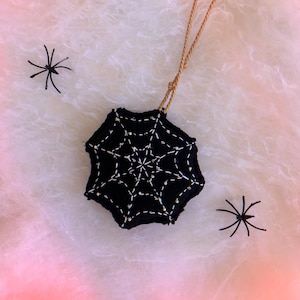 May include: A black fabric ornament shaped like a spider web with white stitched detailing, featuring a gold twisted cord loop for hanging, accompanied by two small black plastic spider figurines on a textured white surface.
