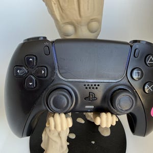 May include: A black video game controller held by a beige, tree-like character figurine. The controller features a central touchpad, directional buttons, two analogue sticks, and the PlayStation logo. The figurine stands on a circular black base.