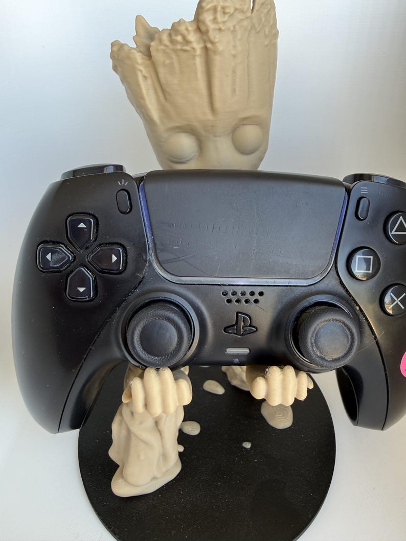 May include: A black video game controller held by a beige, tree-like character figurine. The controller features a central touchpad, directional buttons, two analogue sticks, and the PlayStation logo. The figurine stands on a circular black base.