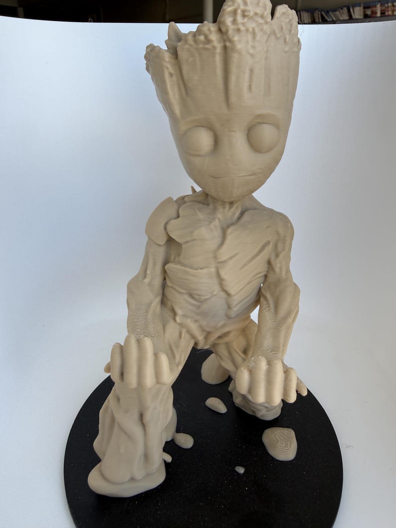 May include: A light beige, 3D-printed figurine of a humanoid tree character with a textured, bark-like surface. The figure has large, round eyes, a small mouth, and is posed in a kneeling position with arms extended forward on a circular black base.