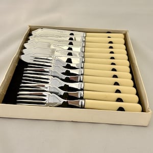 May include: A set of twelve silver-toned metal utensils, including knives and forks, arranged in a box. Each piece features a smooth, cream-colored handle and a polished metal head with decorative engraved detailing near the base of the blade.
