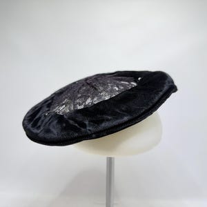 Op de afbeelding: Een zwarte baret met een gladde, effen zwarte fluweelachtige buitenband en een cirkelvormig middenpaneel met een gestructureerd, glinsterend zilver en zwart metallic patroon. De hoed heeft een rond silhouet met een platte bovenkant en zichtbare stiksels.