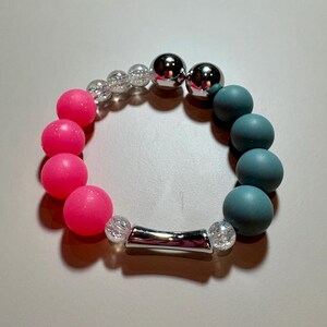 May include: A circular bracelet featuring four vibrant neon pink spherical beads, four matte teal spherical beads, two metallic silver spherical beads, four clear crackle-textured beads, and one central curved silver-toned cylindrical tube bead.