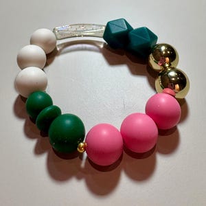 May include: A circular bracelet featuring a variety of beads including matte white spheres, dark green spheres, two dark green geometric shapes, two metallic gold spheres, and three bright pink spheres, connected by a clear glitter-filled tube.