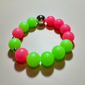 May include: A stretch bracelet featuring alternating neon green and hot pink spherical beads. The design includes two small, reflective silver-toned metallic spherical beads placed at intervals among the colourful matte-finish plastic beads.