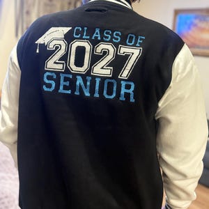May include: A black varsity jacket with white sleeves and striped ribbed trim. The back features white and light blue embroidery that reads "CLASS OF 2027 SENIOR" with a graduation cap icon above the text.