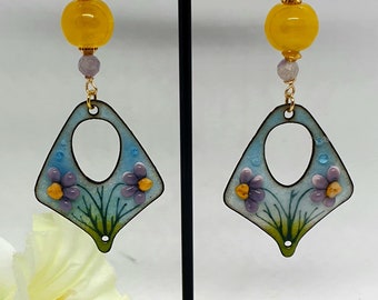 Könnte beinhalten: Ein Paar Hängeohrringe mit einem blauen und grünen Emaille-Design mit lila Blumen. Die Ohrringe sind mit einer gelben Perle und einer goldfarbenen Scheibe verziert.