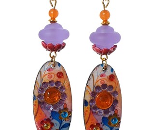 Könnte beinhalten: Hängeohrringe mit orangefarbenen, lila und blauen Blumenmustern. Jeder Ohrring hat eine große orangefarbene Perle oben, die durch goldene Ketten mit einer lila, blütenförmigen Perle und einem ovalen Anhänger mit farbenfrohen Blumenmustern und Edelsteinen verbunden ist.