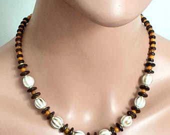 May include: A beaded necklace featuring white, brown, and orange beads. The necklace has a mix of round and disc-shaped beads, with larger white beads interspersed throughout. The necklace is displayed on a mannequin. The text "passioneva créations" is visible.