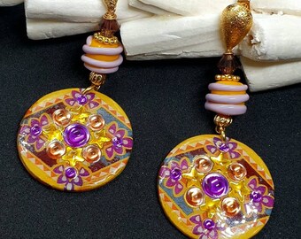 Könnte beinhalten: Ein Paar goldene Ohrringe mit einem runden, gelben, orangefarbenen und violetten Muster. Die Ohrringe haben einen violetten Edelstein in der Mitte des Musters und sind mit goldenen Perlen und einem goldenen tropfenförmigen Anhänger verziert.