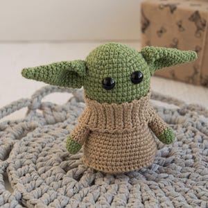 May include: A small, handmade crochet doll featuring a light green head with large, round, black plastic eyes and prominent pointed ears. The doll wears a tan-colored, ribbed-knit sweater that covers its torso and arms, with small green hands visible.
