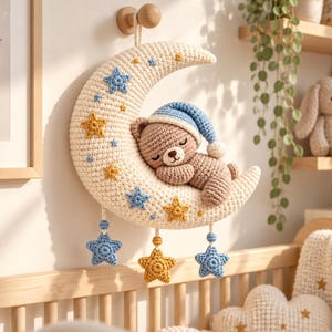 Può includere: Una decorazione da parete all'uncinetto a forma di mezzaluna color crema con un orsacchiotto marrone addormentato che indossa un berretto da notte blu. La luna è decorata con piccole stelle all'uncinetto blu e gialle, con tre stelle pendenti.