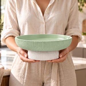 May include: A round, shallow bowl with a light green interior and exterior, featuring a vertical ribbed texture. The bowl is supported by a solid white, circular pedestal base that also displays a matching vertical ribbed pattern.