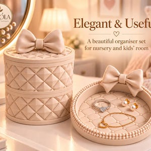 May include: A beige organiser set featuring a cylindrical lidded container and a circular tray, both adorned with a quilted diamond pattern and a decorative bow. The tray holds a silver ring, two pearl earrings, and a gold heart bracelet.