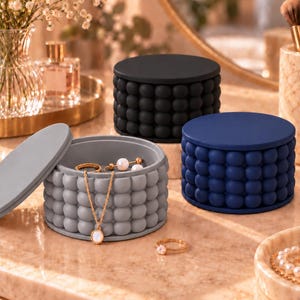 May include: Three round storage containers with textured bubble-patterned sides, available in matte black, navy blue, and light gray. The gray container is open with a matching lid beside it, holding a gold chain necklace and small earrings.