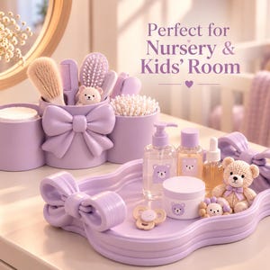 May include: A lavender nursery organizer set featuring a multi-compartment holder with a large bow, containing brushes and cotton swabs. A matching lavender tray with bow handles holds a pump bottle, small jars, a pacifier, and a small teddy bear.