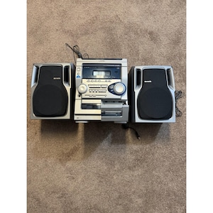 May include: A silver Aiwa stereo system featuring a central unit with a cassette deck, CD player, and radio tuner, flanked by two matching silver and black speakers. The central console includes various control buttons and two circular dials.