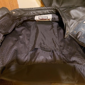 May include: A black leather jacket with a mesh interior lining and a Firstgear brand label at the neck. The jacket features a central pocket with a velcro closure, metal snap buttons on the collar, and textured blue fabric accents.