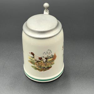May include: A cream-colored ceramic beer stein featuring a detailed illustration of a brown and white hunting dog in a grassy field. The stein has a smooth, matte-finish metal lid with a small spherical knob and a thin green band near the base.