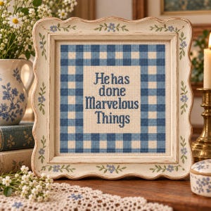 Könnte beinhalten: Ein quadratisches Kreuzstich-Stickbild in einem cremefarbenen Holzrahmen mit gewelltem Rand und blauen Blumenmotiven. Die Stickerei zeigt einen blau-weißen Vichy-Karo-Rahmen, der den Text "He has done Marvelous Things" in blauer Schrift umschließt.