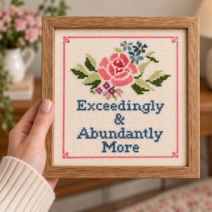 Può includere: Un ricamo a punto croce quadrato in una cornice di legno chiaro con una rosa rosa, fiori blu e viola e foglie verdi. Il testo "Exceedingly & Abundantly More" è ricamato in lettere blu, il tutto racchiuso all'interno di un bordo rosa.