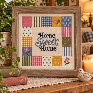 Pode incluir: Um bordado em ponto cruz quadrado numa moldura de madeira clara com o texto Home Sweet Home em azul centrado dentro de uma borda de quadrados de patchwork coloridos incluindo bolinhas, riscas, xadrez e pequenos motivos florais em tecido bege.