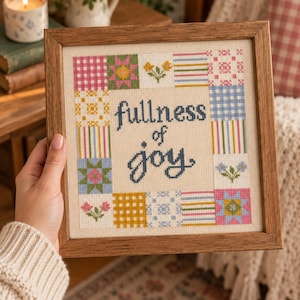 Op de afbeelding: Een vierkant kruissteek borduurwerk in een houten lijst met de tekst fullness of joy in donkerblauw schrift. De centrale tekst is omringd door een rand van kleurrijke patchwork-stijl vierkanten met bloemen-, ruit- en streepmotieven.