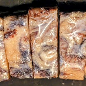 May include: Five rectangular cuts of meat, individually wrapped in clear plastic. The meat has a marbled appearance with shades of red, brown, and cream. The cuts are arranged side by side on a dark surface.