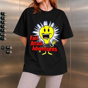 Puede incluir: Una camiseta negra de manga corta con un gráfico de una bombilla amarilla de dibujos animados con una amplia sonrisa y una mano saludando. El texto "Fact Attack Adventures" está impreso en letras rojas negritas junto al personaje sobre un fondo blanco.