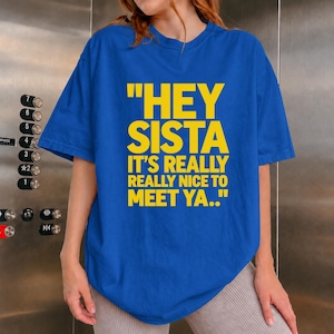Puede incluir: Una camiseta azul brillante de manga corta con texto amarillo llamativo que dice: "HEY SISTA IT'S REALLY REALLY NICE TO MEET YA.." La prenda tiene un cuello redondo y un corte holgado, mostrando el estampado gráfico claramente en el pecho.