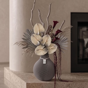 Könnte beinhalten: Eine graue, spiralförmig gerippte Vase enthält ein Blumenarrangement aus cremefarbenen Anthurien mit dunklen Spitzen, tiefroten Calla-Lilien, getrockneten Palmblättern, hohen dünnen Zweigen und langen, herabhängenden dunkelroten Amaranthfäden.