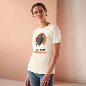 May include: A cream-colored short-sleeve t-shirt featuring a graphic of a person's profile with floral hair against an orange circle. Below the graphic, the text reads "SOFT HEART. HARD BOUNDARIES." in black and orange capital letters.