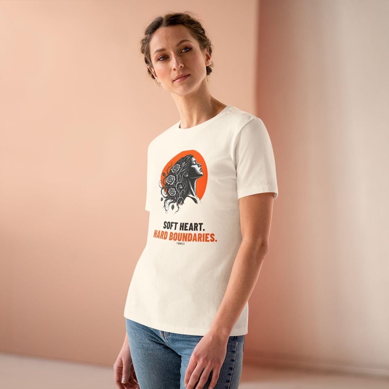 May include: A cream-colored short-sleeve t-shirt featuring a graphic of a person's profile with floral hair against an orange circle. Below the graphic, the text reads "SOFT HEART. HARD BOUNDARIES." in black and orange capital letters.