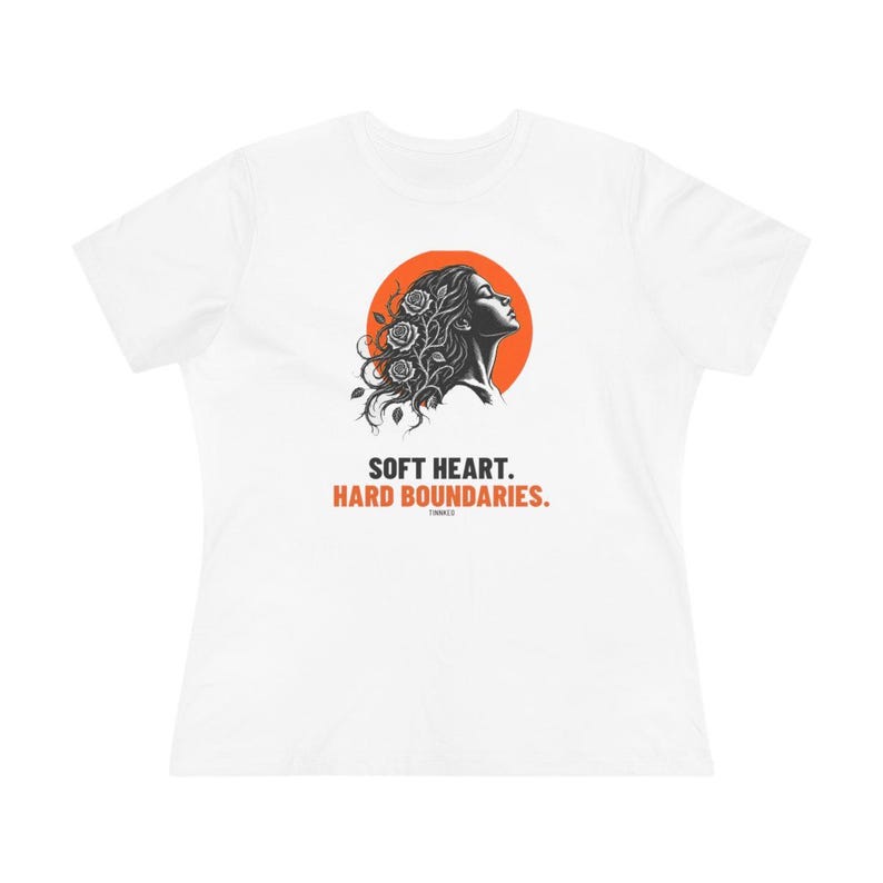 May include: A white short-sleeve t-shirt featuring a graphic of a person in profile with roses in their hair against an orange circle. Below the graphic, the text reads: SOFT HEART. HARD BOUNDARIES. The shirt has a crew neckline and a standard fit.
